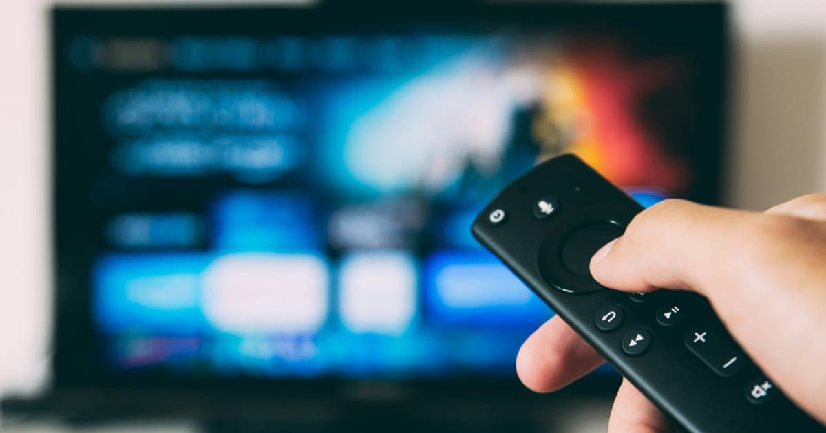 Hand holding a Fire TV-style voice remote pointed at a television with a blurred blue streaming interface