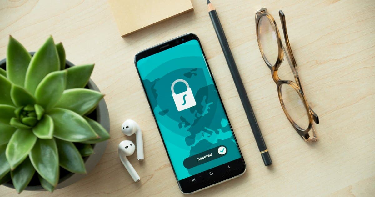 Smartphone on a desk showing a padlock security screen marked Secured, next to earbuds, a pencil and glasses
