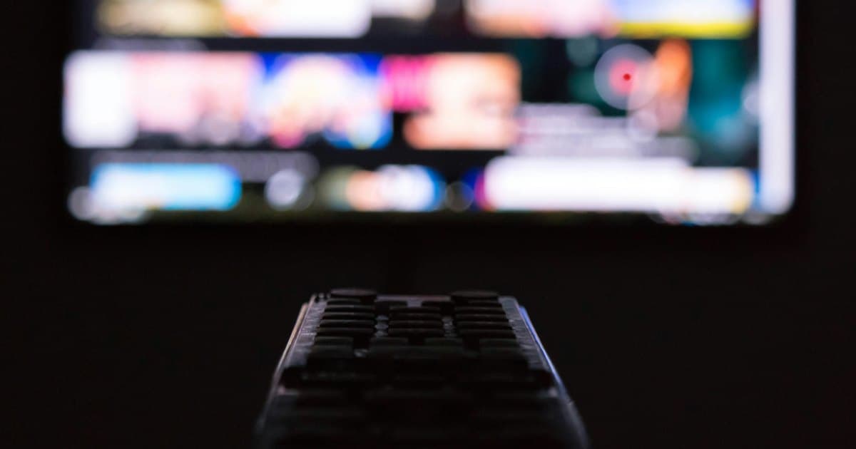 TV remote control in a dark room, pointed at a television showing a blurred grid of streaming content